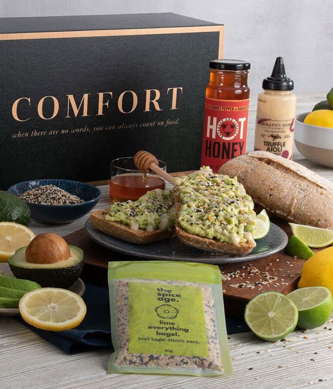Central is a plate of avocado toast with a bag of lime everything bagel seasoning, lime & lemon slices, a loaf of bread, jar of honey, bowl of everything seasoning, and sliced avocados appearing around the plate.