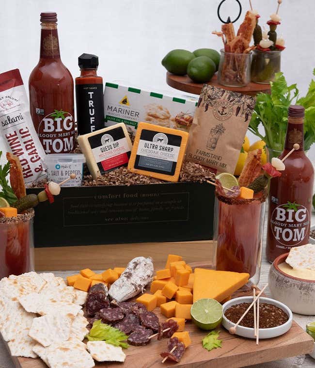 A gift box with blocks of queso &amp; cheddar cheese, packet and bottle of bloody mary mix, salt &amp; pepper seasoning blend, box of water crackers, hot sauce from Truff. In front of the box is a grazing board with crackers, sliced soppressata, cubed cheddar, an