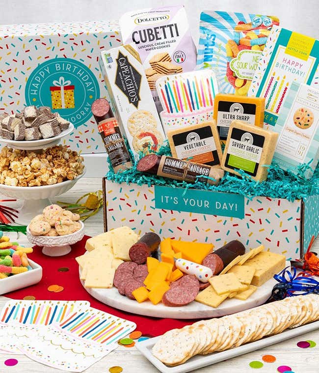 In a gift box, a bag of of chocolate wafter Cubetti's, a box of crackers, a bag of gummy worms, a box of kettle corn, three blocks of cheese, two packages of sausage. In front of the box, a plate of sliced meat &amp; cheese and a plate of crackers. Beside the