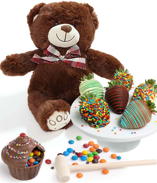 A teddy bear plush next to a breakable chocolate cupcake with a wooden mallet on a wooden serving tray, and 6 chocolate dipped strawberries with drizzles and sprinkles