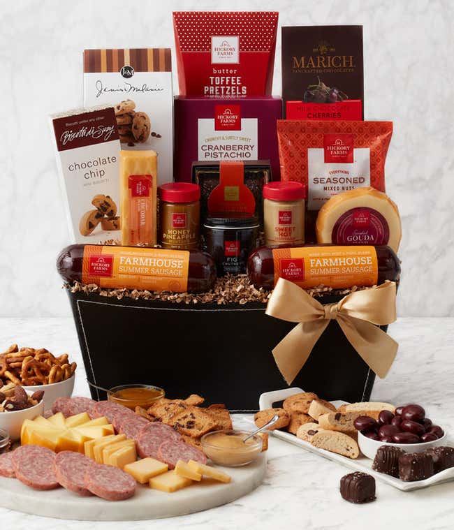 A  black gift basket with two packs of sausage, a wheel of gouda cheese, a block of cheddar cheese, a jar of honey pineapple mustard, a jar of hot &amp; sweet mustard, and fig chutney, a box of chocolate chip mini biscotti, chocolate chip cookies, toffee pret