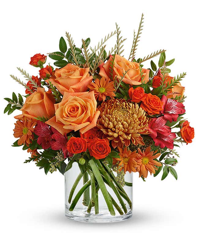 A vibrant floral arrangement featuring orange roses, orange spray roses, red Peruvian lilies, bronze mums, and orange daisies, all elegantly displayed in a clear cylinder vase.