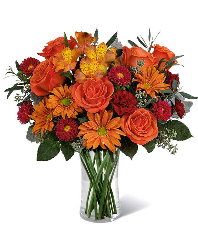 A vibrant floral arrangement featuring orange roses, orange alstroemeria, bronze daisies, red Matsumoto asters, maroon mini carnations, and seasonal greenery, complemented by a personal card message, all beautifully presented in a clear cylinder vase.