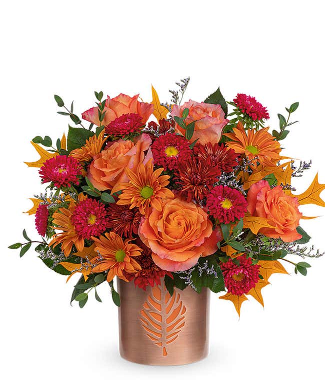 A vibrant floral arrangement featuring orange roses, red Matsumoto asters, bronze cushion spray chrysanthemums, bronze daisy spray chrysanthemums, purple limonium, parvifolia eucalyptus, and yellow oak leaves, all elegantly arranged in a bronze vase.